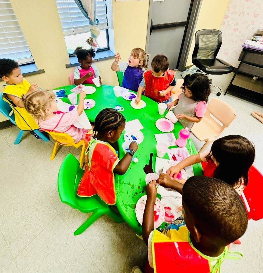 Toddlers in colorful smocks painting paper plates at a round green table